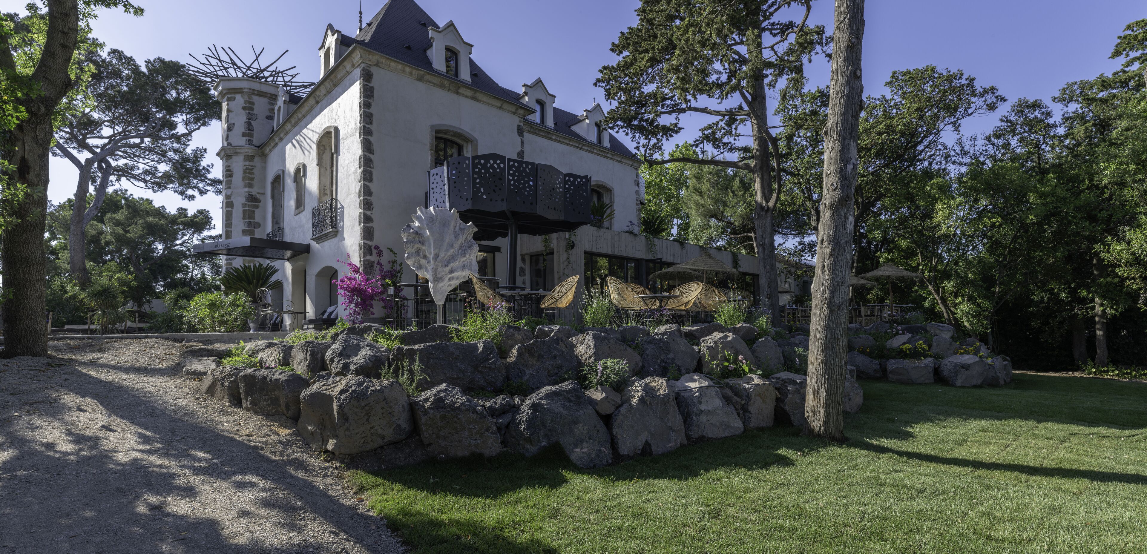 A large, historic building with a stone facade, a balcony with latticework, and a well-maintained garden with rocks and trees.