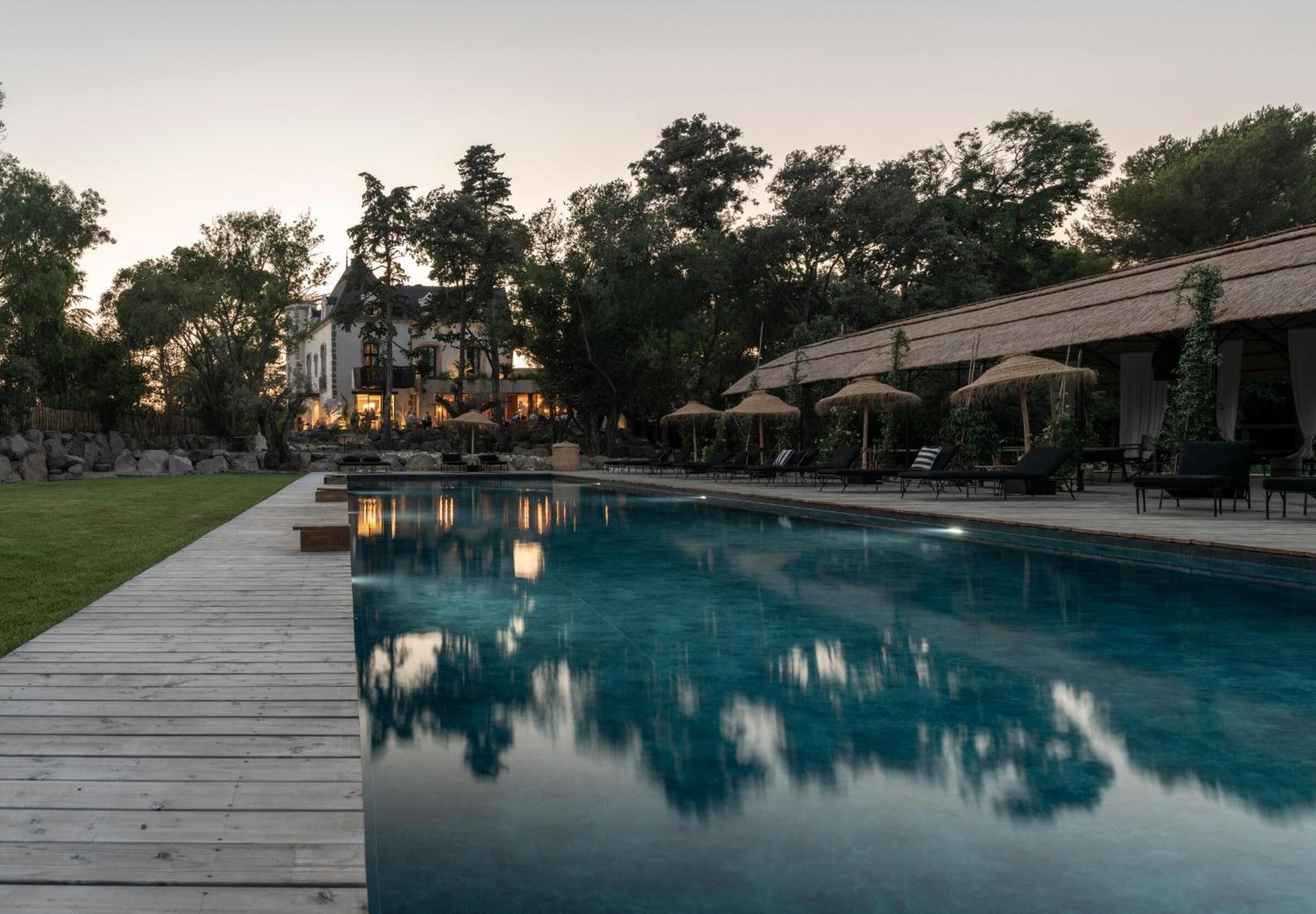 Una piscina con sillas de descanso y sombrillas, rodeada de árboles y un edificio al fondo.