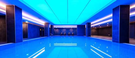A large indoor swimming pool with a modern design, featuring a central seating area and a blue-lit ceiling.
