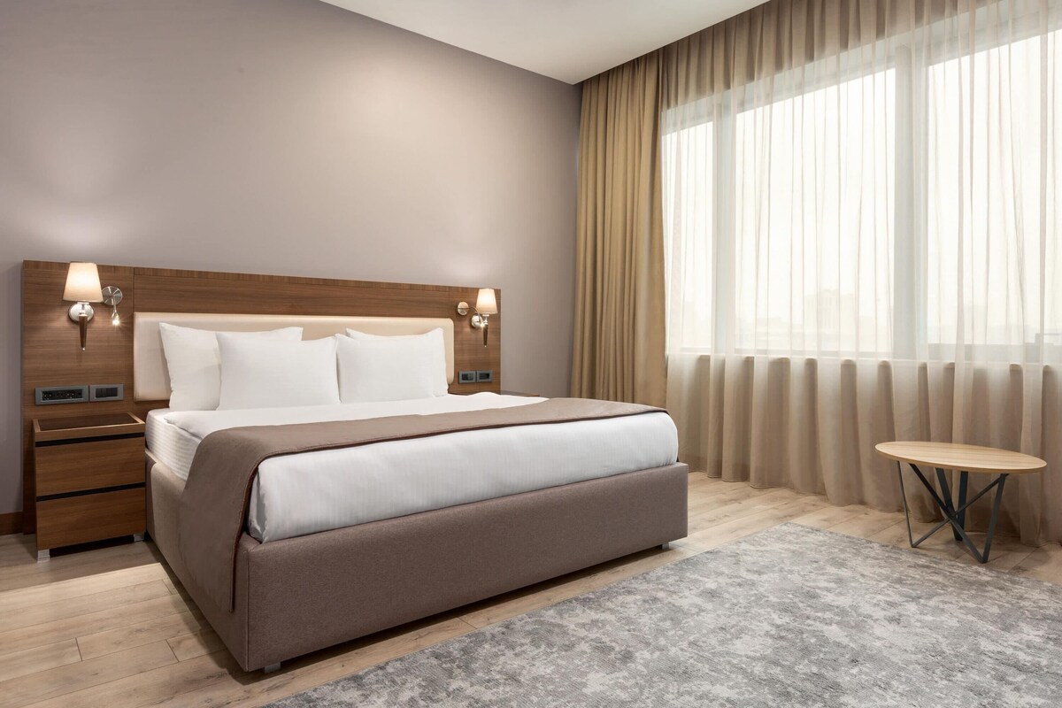 A hotel room with a large bed, bedside tables, a small round table, and sheer curtains.