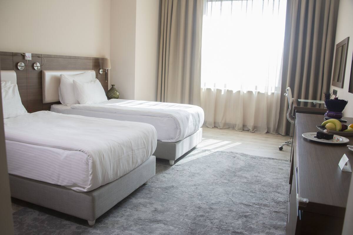 A hotel room with two beds, a desk with a chair, a lamp, and a window with curtains.