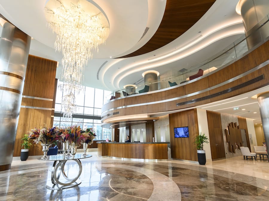 A modern hotel lobby with a reception desk, a chandelier, and a large plant.