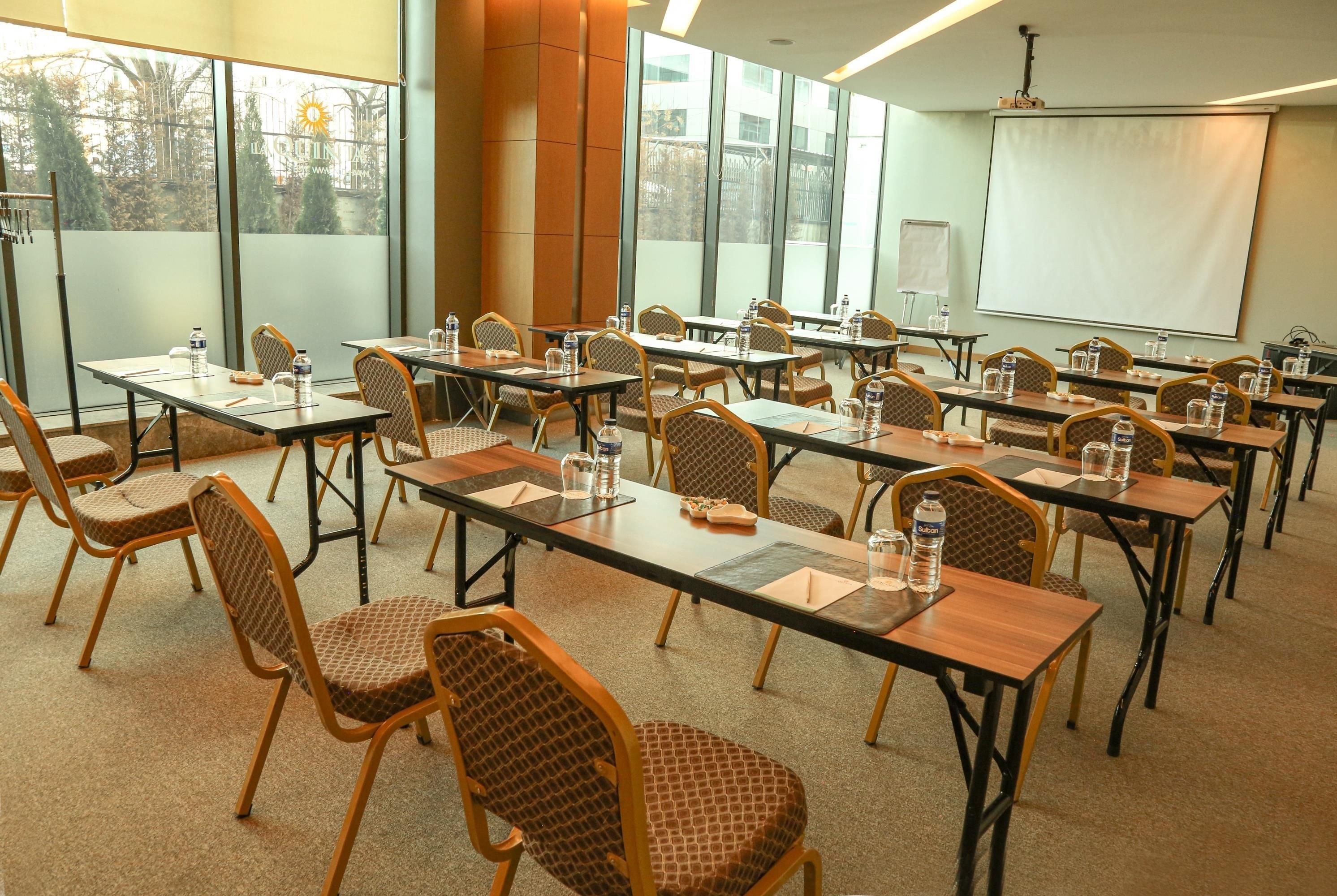 A conference room with multiple tables and chairs, large windows, and a projector screen.