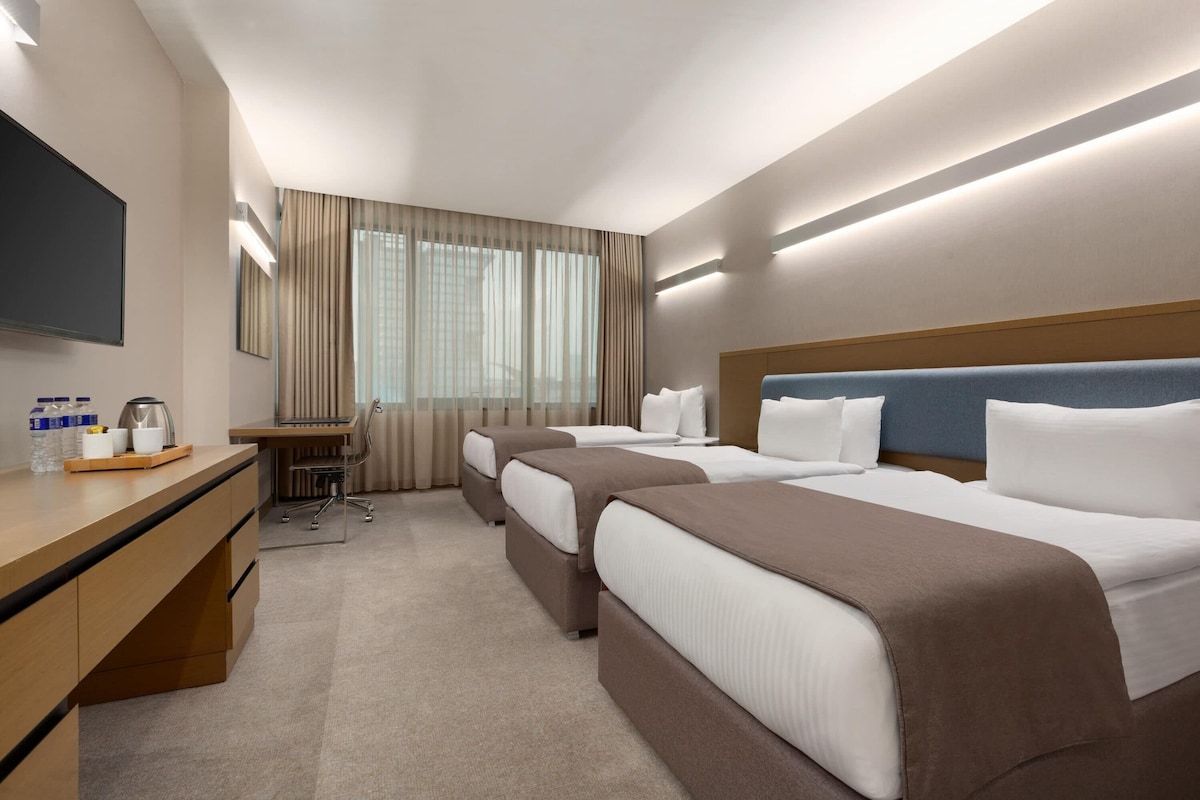 A hotel room with two beds, a desk, a flat-screen TV, and a large window with curtains.