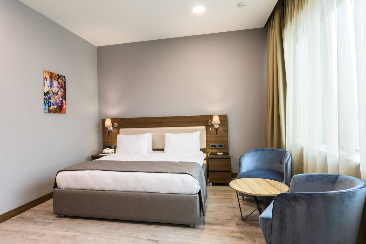 A modern hotel room with a large bed, a bedside table, a chair, a small round table, and a painting on the wall.