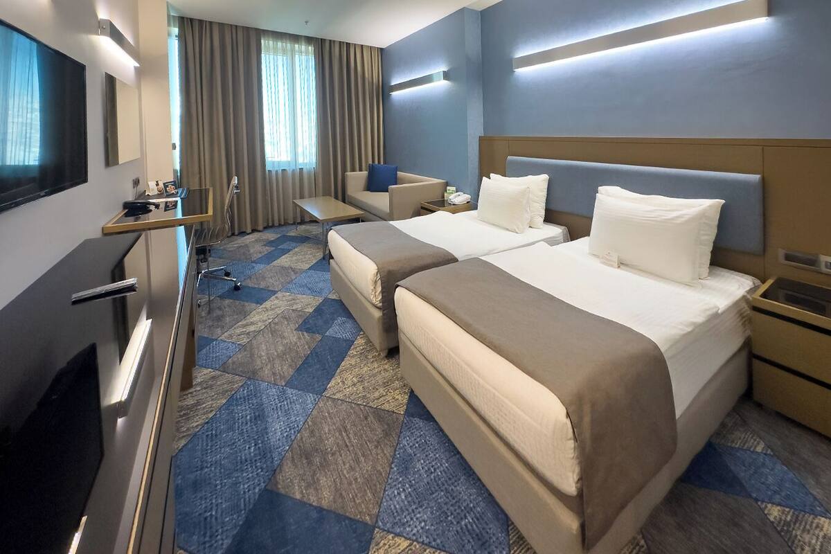 A hotel room with two beds, a desk, a TV, and a large window with curtains.
