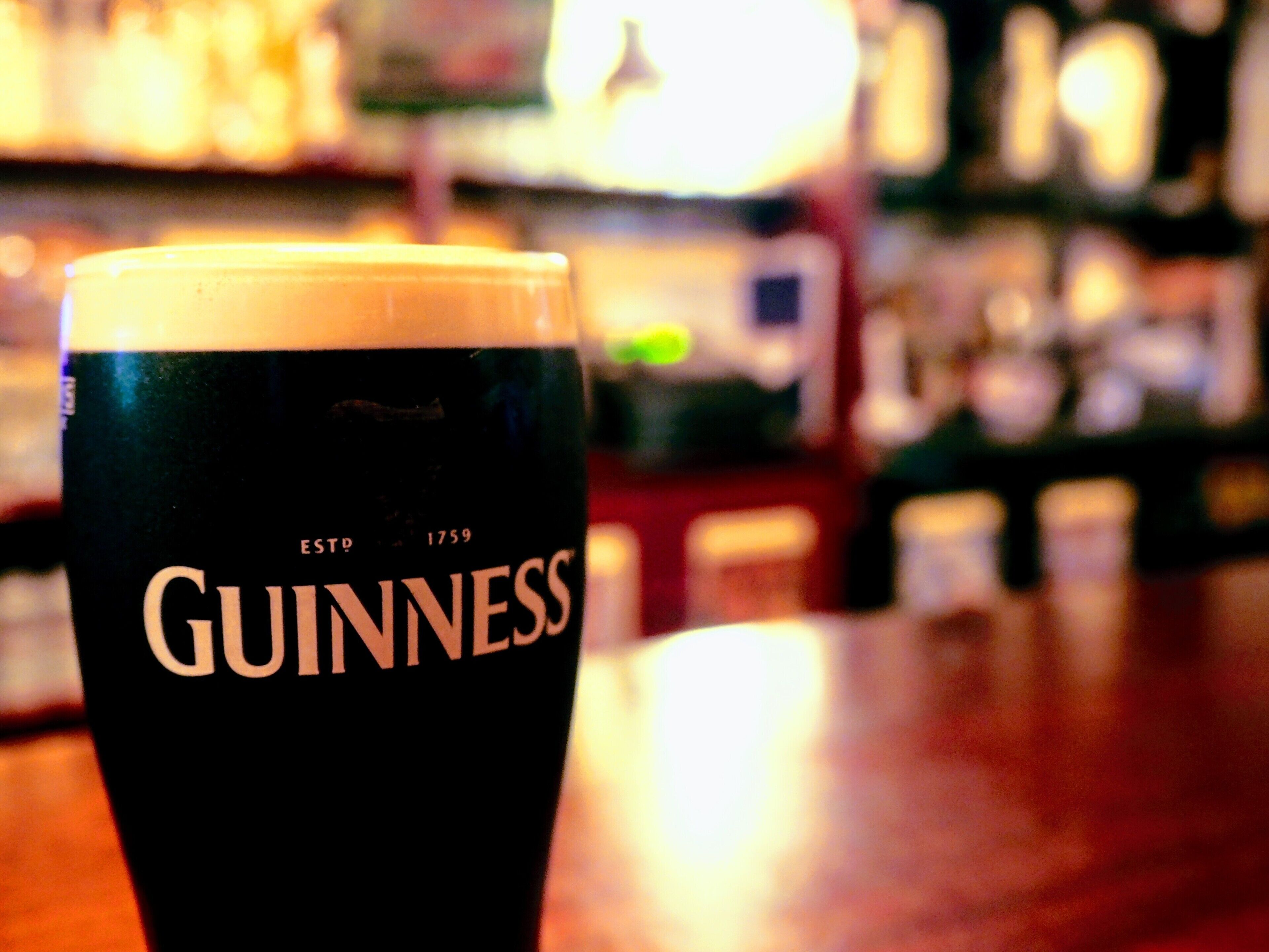 A close-up of a Guinness beer glass on a bar counter with a blurred background of shelves and bottles.