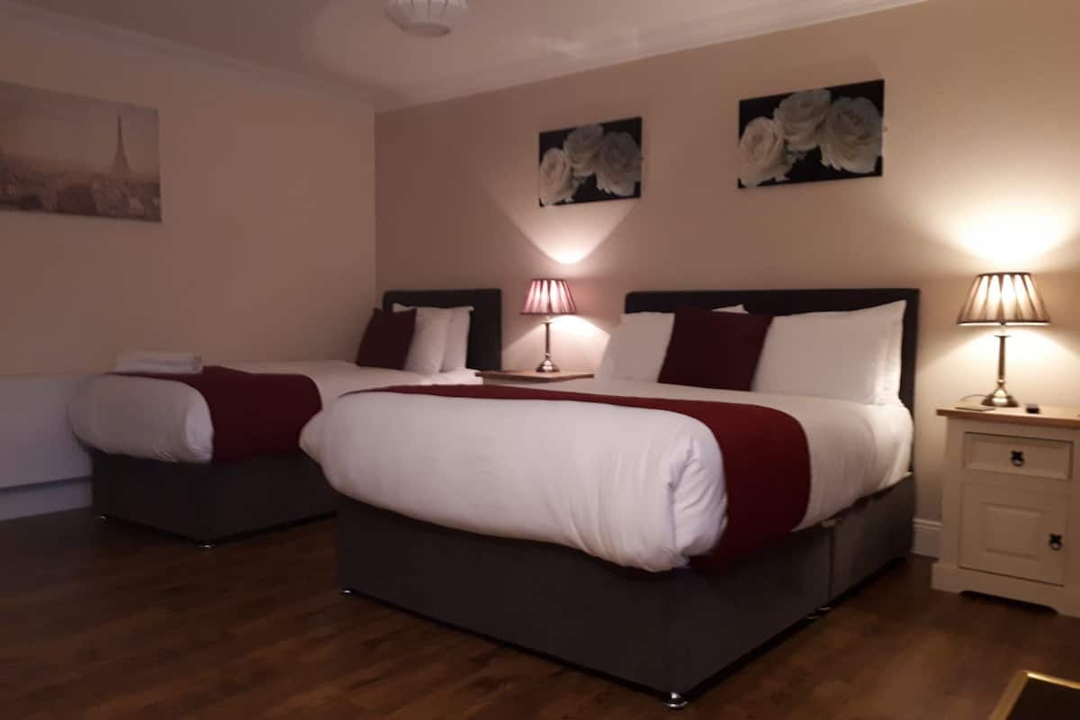 A hotel room with two beds, each with a red throw blanket and white bedding. There are two bedside tables with lamps, and the walls are decorated with floral paintings.