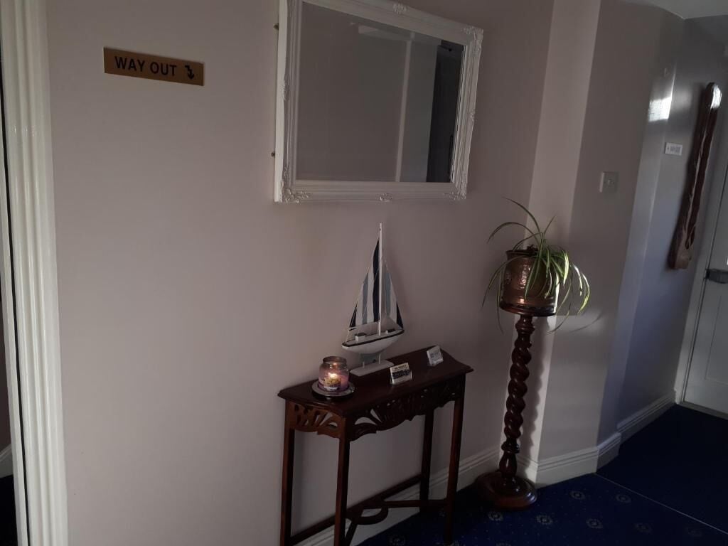 A hallway with a 'WAY OUT' sign, a mirror, a small table with a candle and a ship model, and a potted plant.