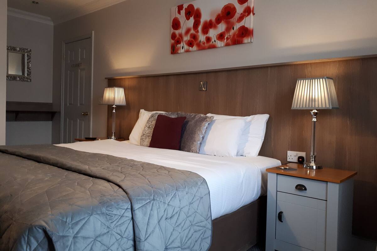 A neatly made bed with a grey quilt, two bedside lamps, a wooden headboard, and a red poppy artwork on the wall.