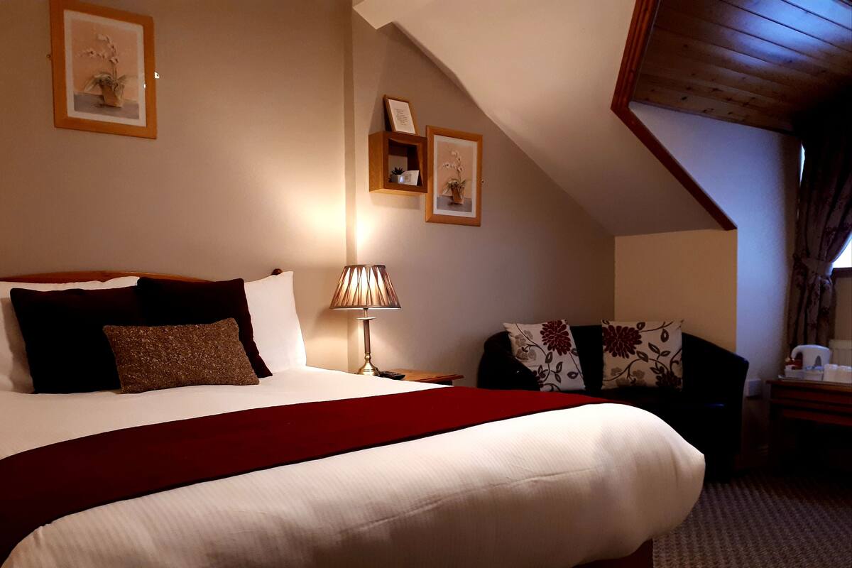 A bedroom with a large bed, a bedside table with a lamp, a chair with floral cushions, and framed pictures on the walls.