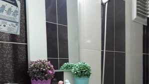 Un baño con un lavamanos blanco, un espejo, una ducha y dos plantas en macetas sobre una estantería.