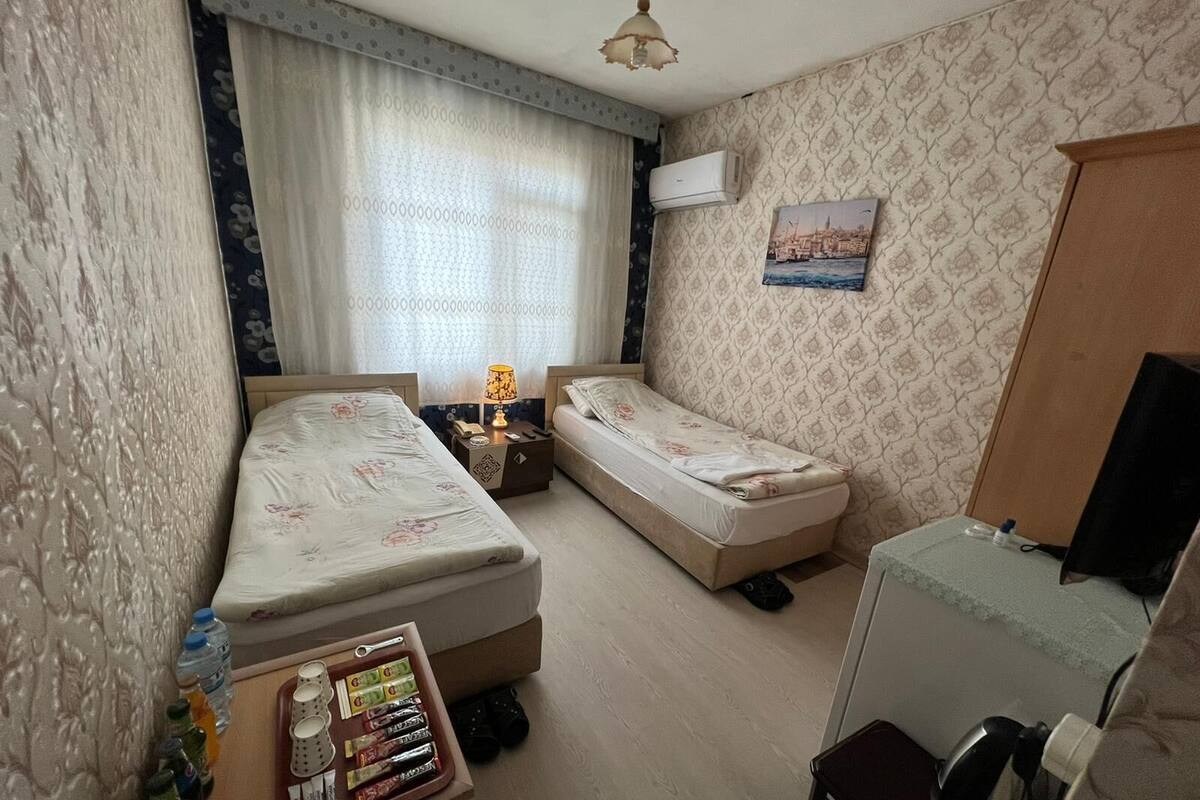 A hotel room with two single beds, a small desk with a chair, a nightstand with a lamp, a wall-mounted air conditioner, and a picture of a cityscape.