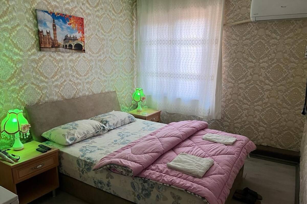 A bedroom with a bed, a nightstand, a lamp, a picture of a cityscape, and a window with curtains.
