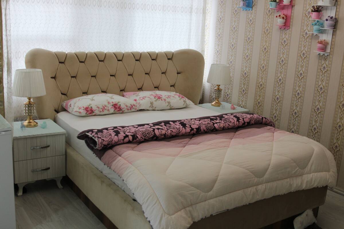 A bedroom with a tufted headboard, two bedside tables with lamps, a floral bedspread, and a patterned wall.
