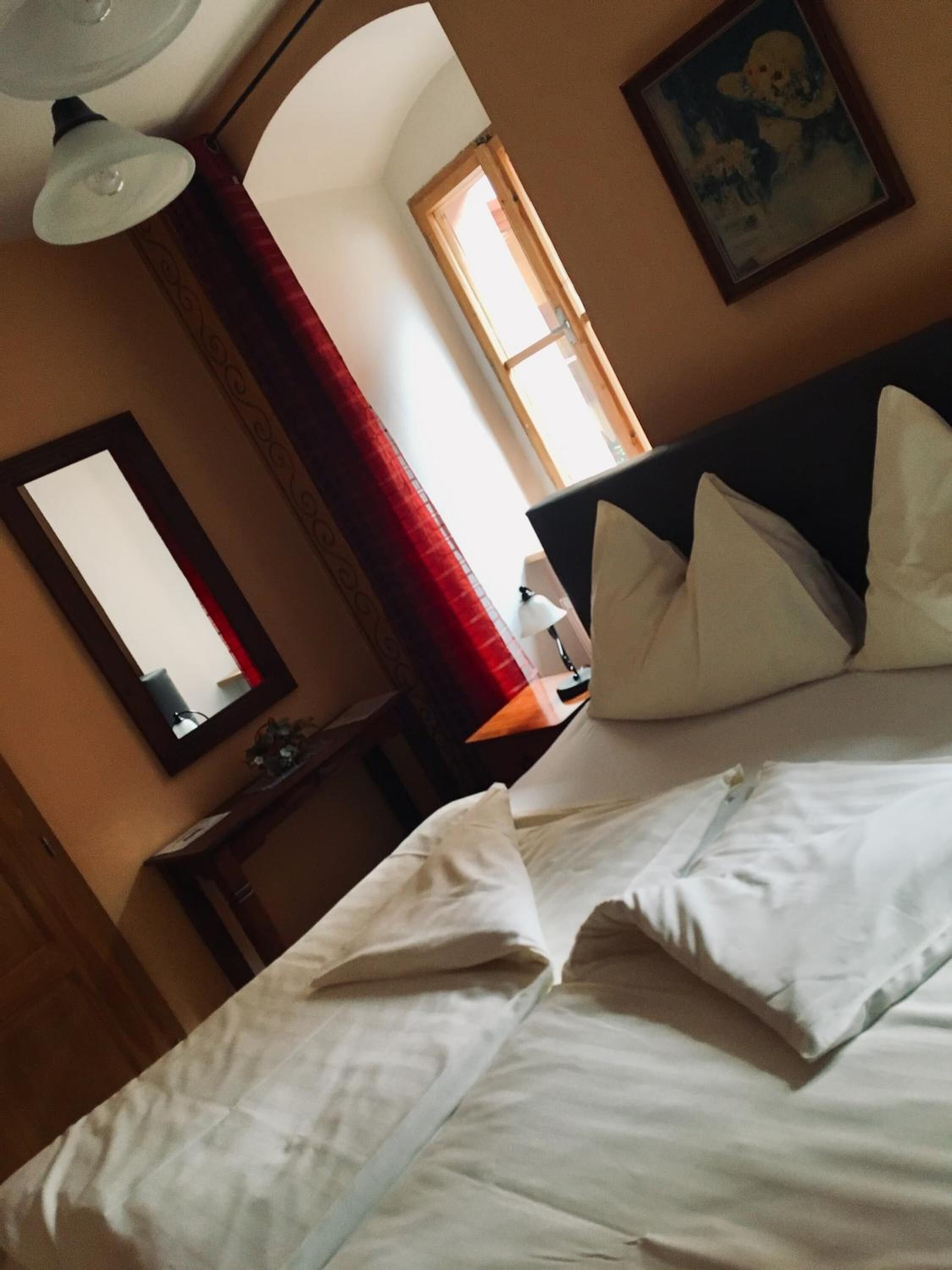A hotel room with a bed, a mirror, a window with red curtains, and a painting on the wall.