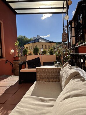A covered outdoor seating area with a wicker sofa and a small table, overlooking a building with a blue roof. - Atmosfera (Loket)