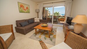 A living room with a sofa, a coffee table, and a TV. There is a view of the ocean through the glass doors.