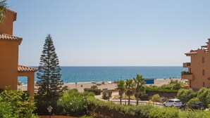 A coastal view with a beach, palm trees, and buildings with Mediterranean architecture.