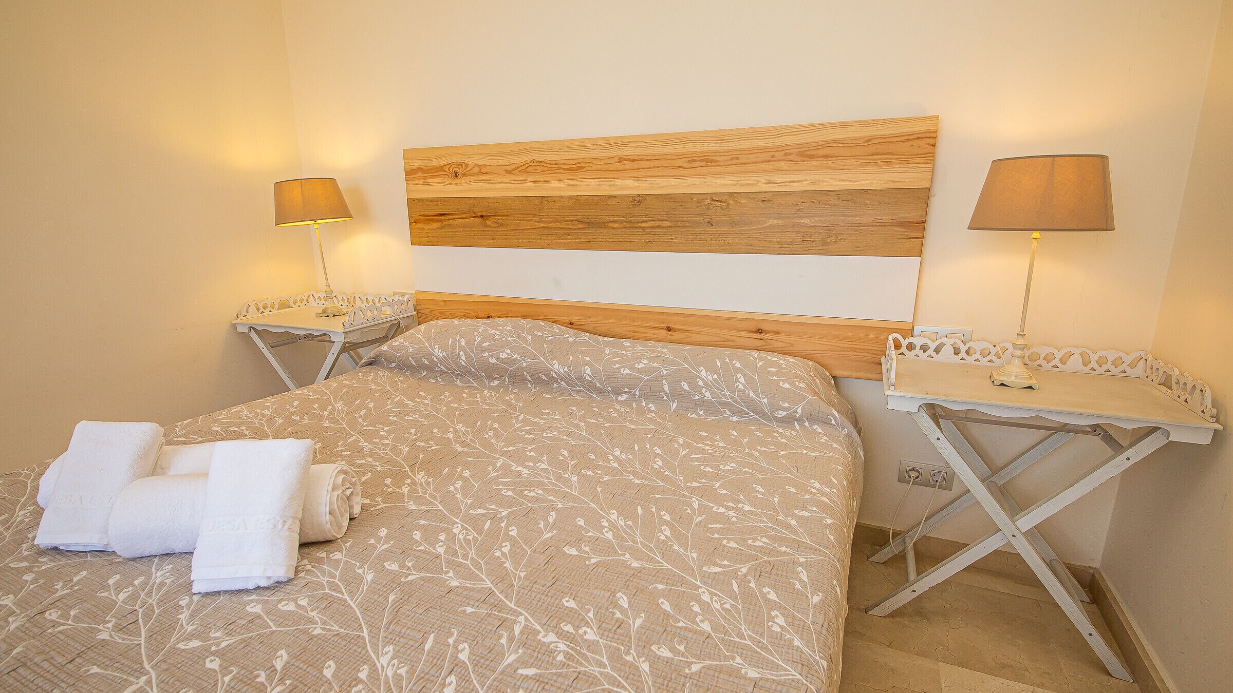 Un dormitorio con cabecera de madera, dos mesitas de noche con lámparas, una cama con colcha estampada y dos toallas enrolladas sobre la cama.