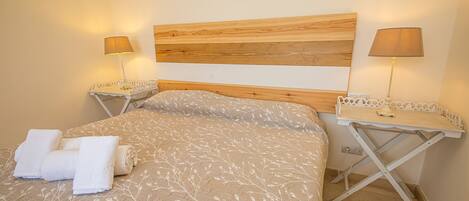 Un dormitorio con cabecera de madera, dos mesitas de noche con lámparas, una cama con colcha estampada y dos toallas enrolladas sobre la cama.