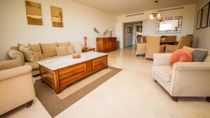 A spacious living room with a large wooden coffee table, beige sofas, and a dining area with wicker chairs.