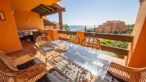 A glass-top table with wicker chairs, a small sofa, and a view of the ocean and buildings in the background.