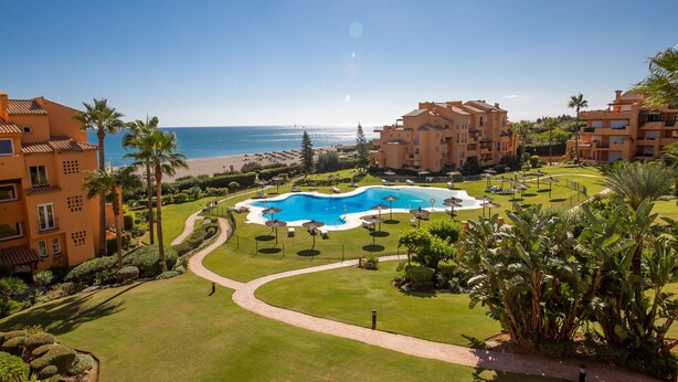 A resort with a central swimming pool, multiple buildings, and a beach view.