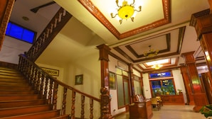Una imponente escalera de madera con barandillas y un candelabro, que conduce a una zona de recepción con un mostrador y sillas.