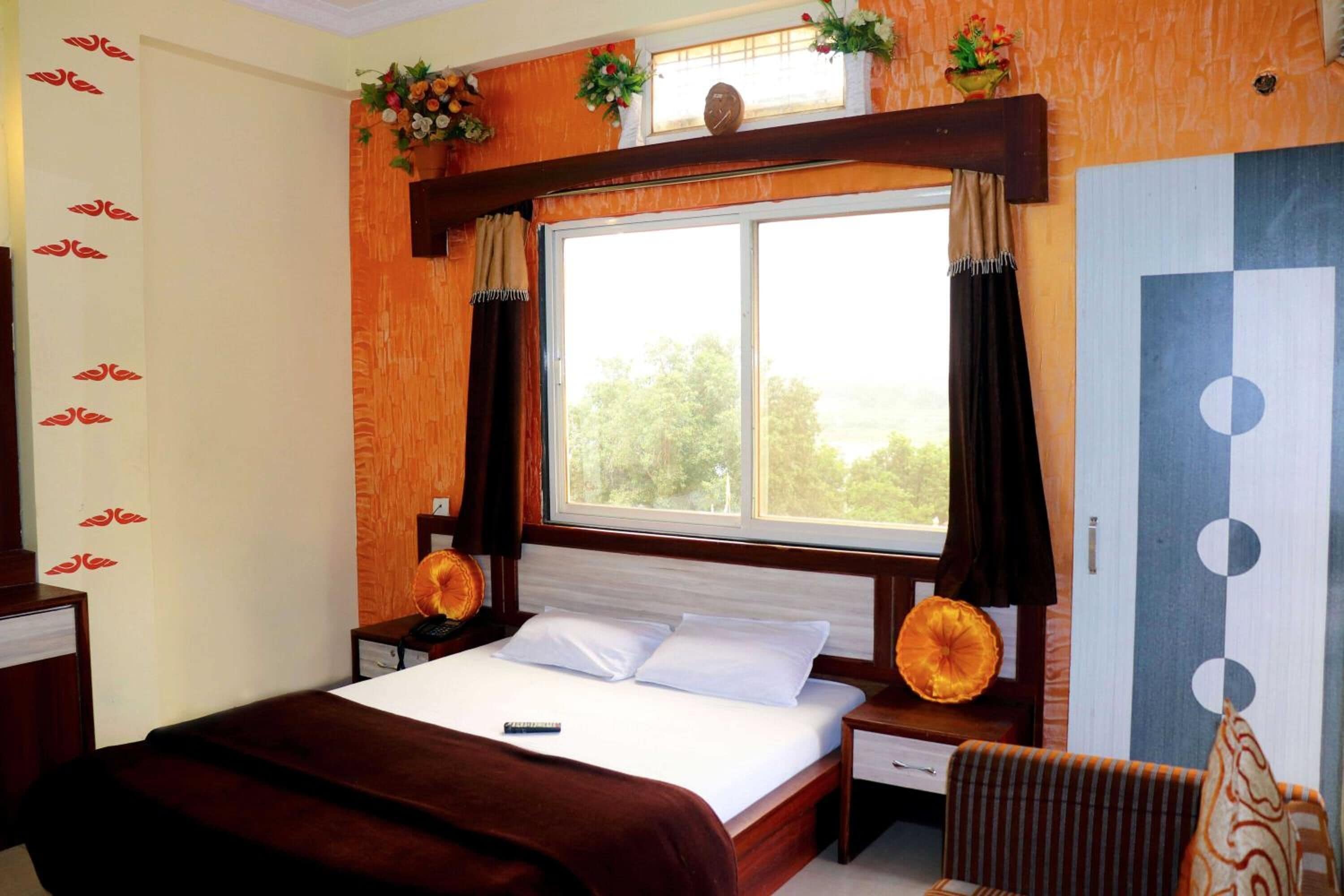 A bedroom with a large window, a bed with white linens, a wooden headboard, and a nightstand with a lamp.