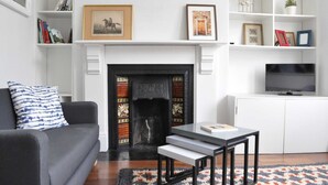 Een woonkamer met een open haard, een bank, een bijzettafeltje, een televisie en planken met boeken en decoraties.
