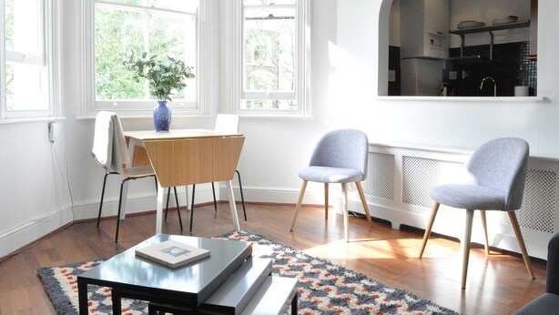 Een lichte kamer met een houten eettafel, twee stoelen, een klein bijzettafeltje en een uitzicht op een keuken via een boogvormige doorgang.