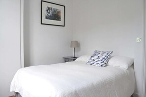 A neatly made bed with white bedding and a blue patterned pillow, a small bedside table with a lamp, and a framed picture on the wall.