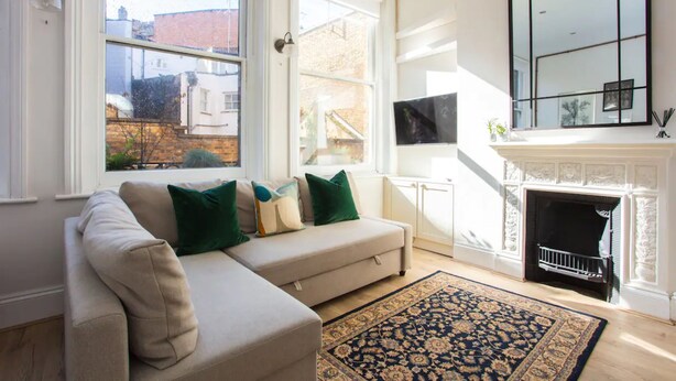 A living room with a beige sofa, green cushions, a patterned rug, a fireplace, and a large window with a view of buildings outside. - Stylish Apartment in Central London - Farringdon (London)