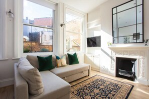 A living room with a beige sofa, green cushions, a patterned rug, a fireplace, and a large window with a view of buildings outside. - Stylish Apartment in Central London - Farringdon (London)