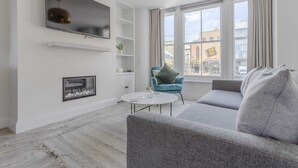 Een moderne woonkamer met een grijze bank, een blauwe fauteuil, een flatscreen televisie aan de muur, een klein wit bijzettafeltje en een open haard.