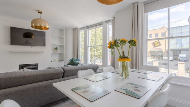 Een moderne woonkamer met een witte eettafel, een grijze bank en een groot raam met uitzicht op de straat.