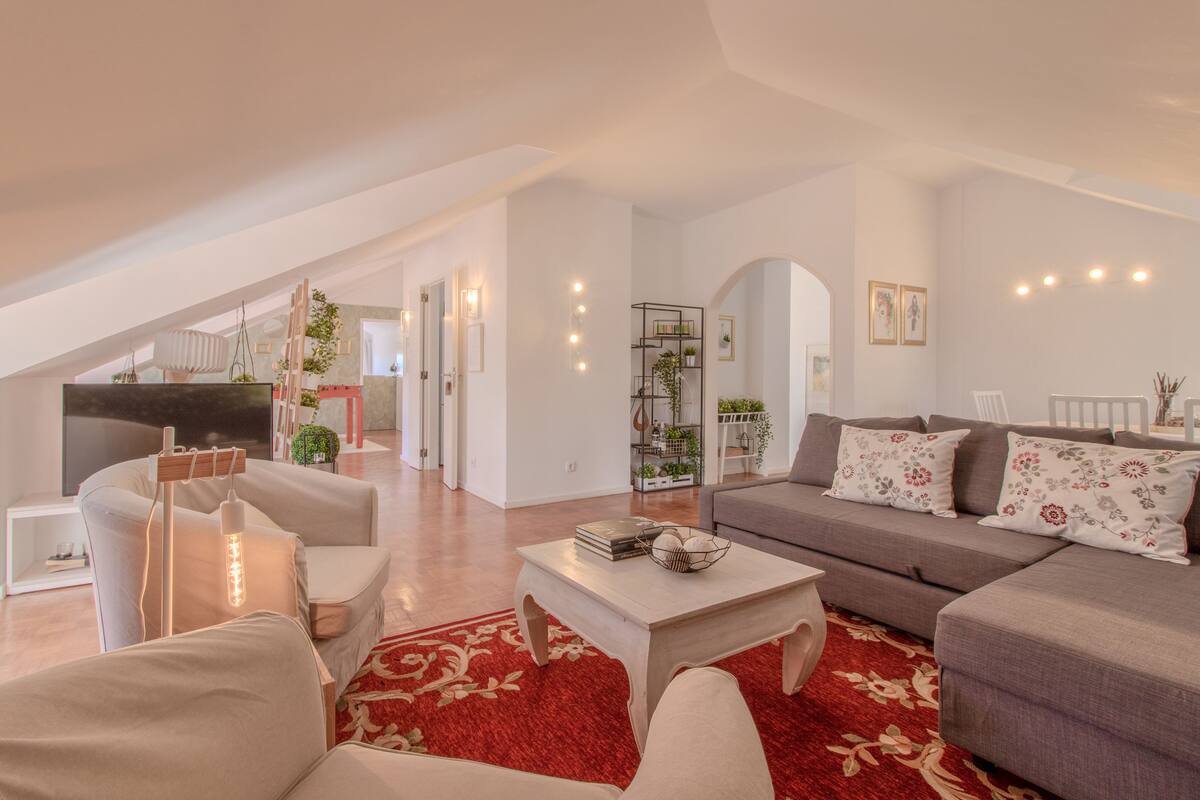 A spacious living area with a high ceiling, a red patterned rug, and a grey sofa with floral cushions.