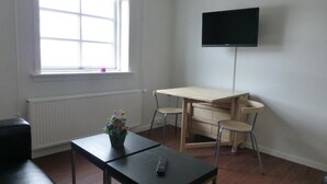 A room with a wooden table, two chairs, a TV on the wall, a window with blinds, and a radiator. - IceCap Apartments (Ilulissat)