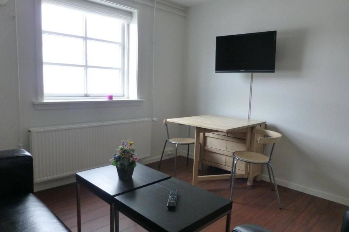 A room with a wooden table, two chairs, a TV on the wall, a window with blinds, and a radiator.