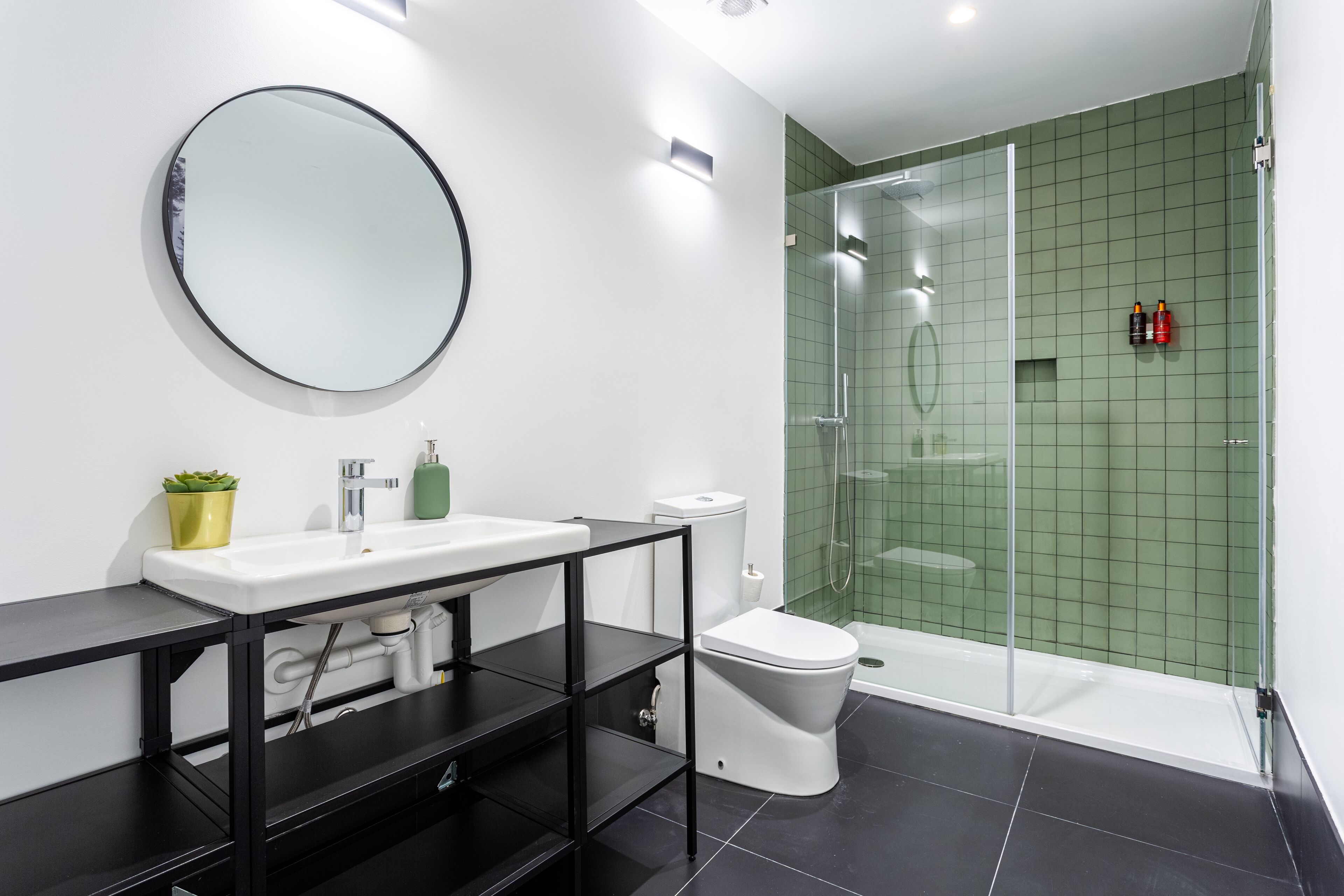 A modern bathroom with a white sink, black shelves, a round mirror, a toilet, and a glass-enclosed shower.