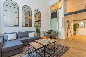 A modern living room with a grey sectional sofa, a wooden coffee table, and a colorful rug. There are three arched mirrors on the wall and a large window with a view of the interior. - Bairro Alto Bronze of Art Apartments (Lisbon)