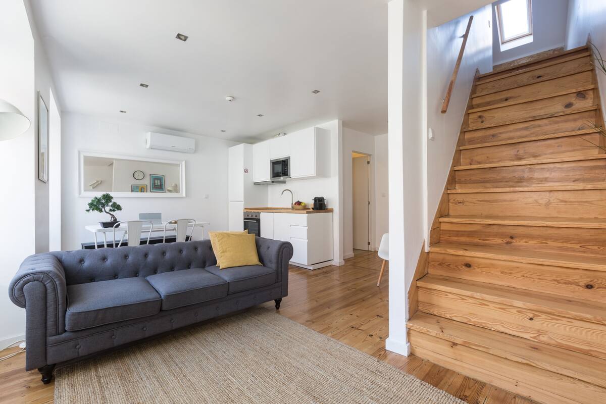 A modern living space with a grey sofa, wooden staircase, white kitchen, and a dining area with a table and chairs.