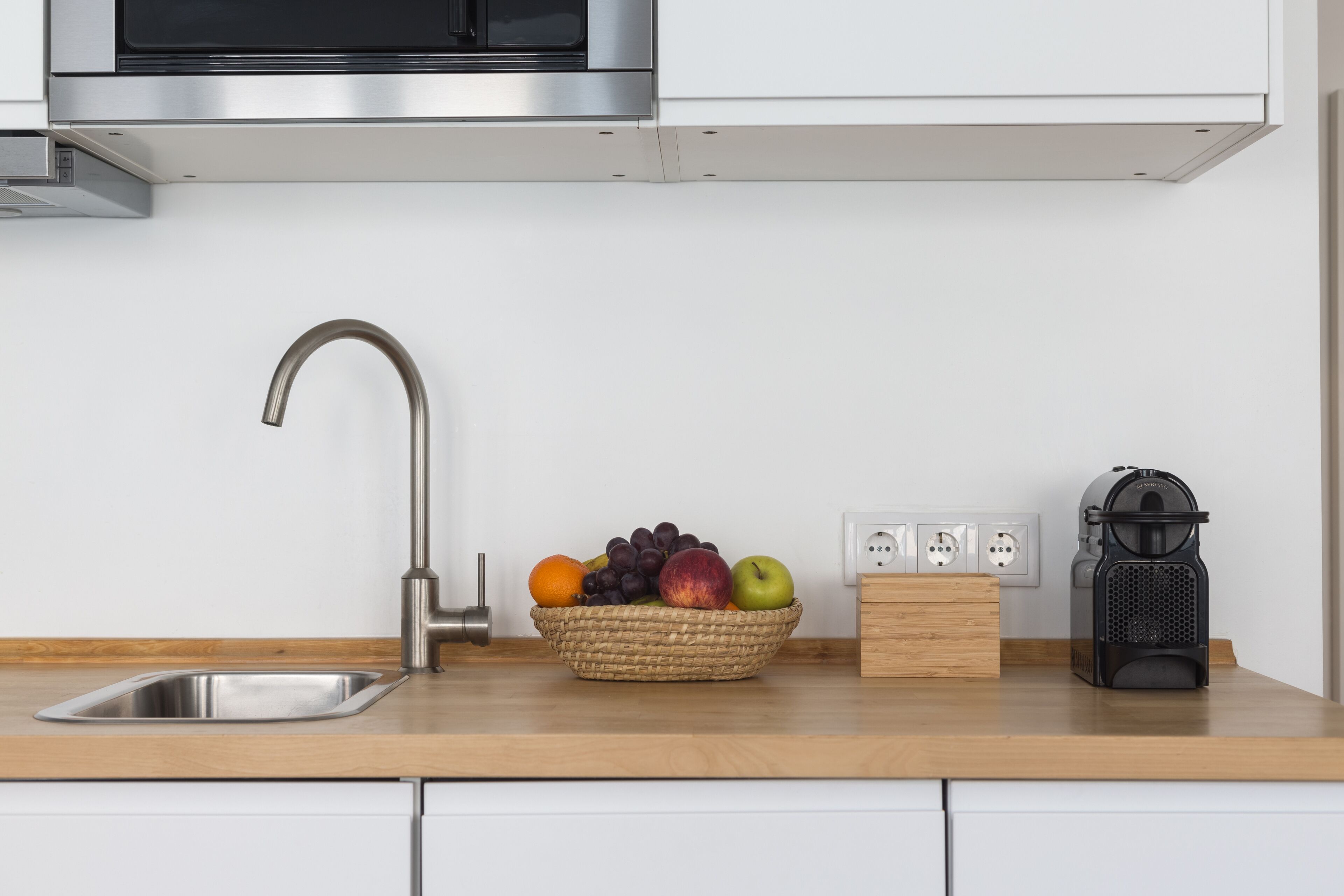 Une cuisine équipée d’un évier en acier inoxydable, d’un panier de fruits, d’une machine à café et d’un four à micro-ondes placé au-dessus du plan de travail.