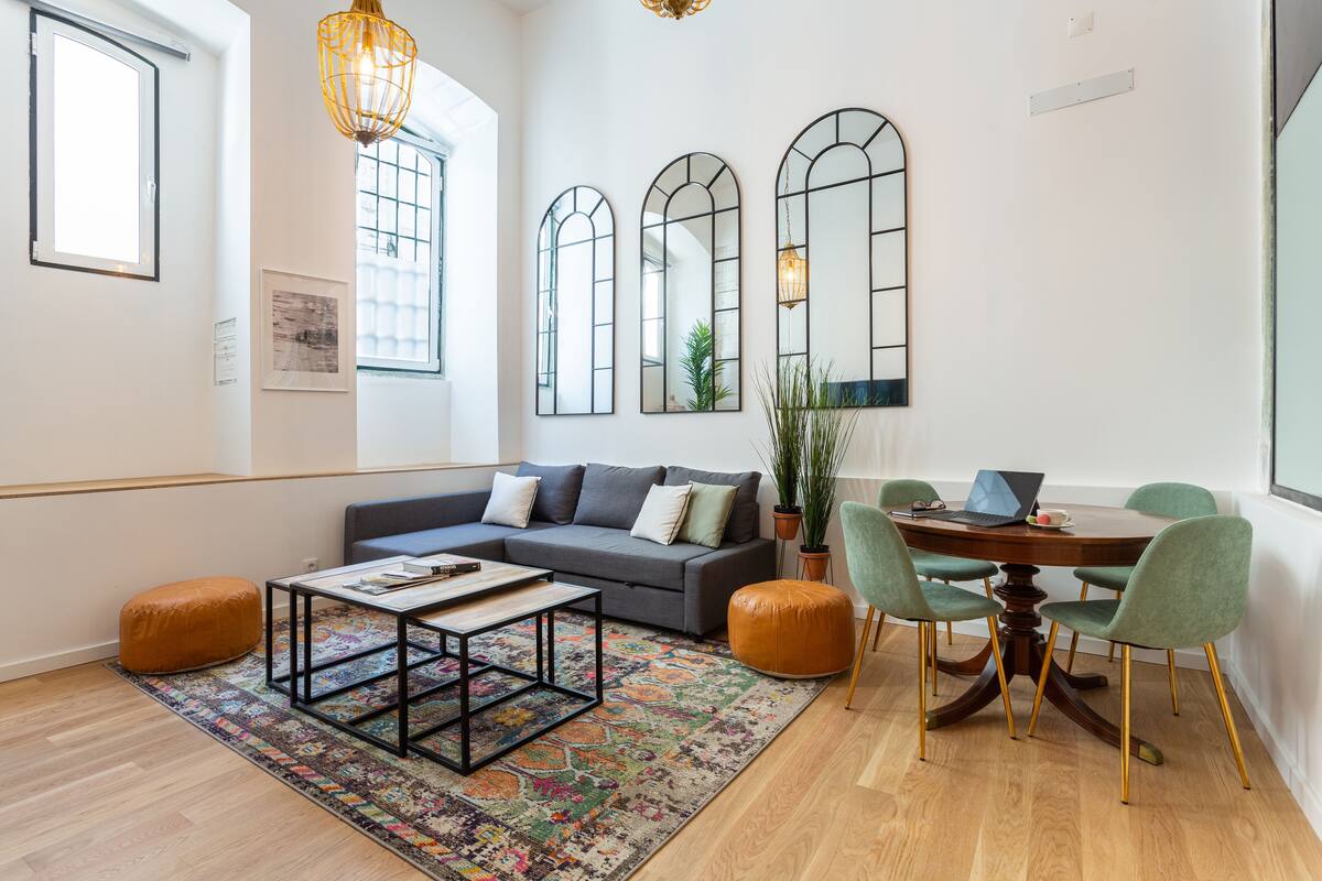 A modern living room with a grey sofa, a coffee table, and a dining table with chairs. There are large windows and mirrors on the walls.