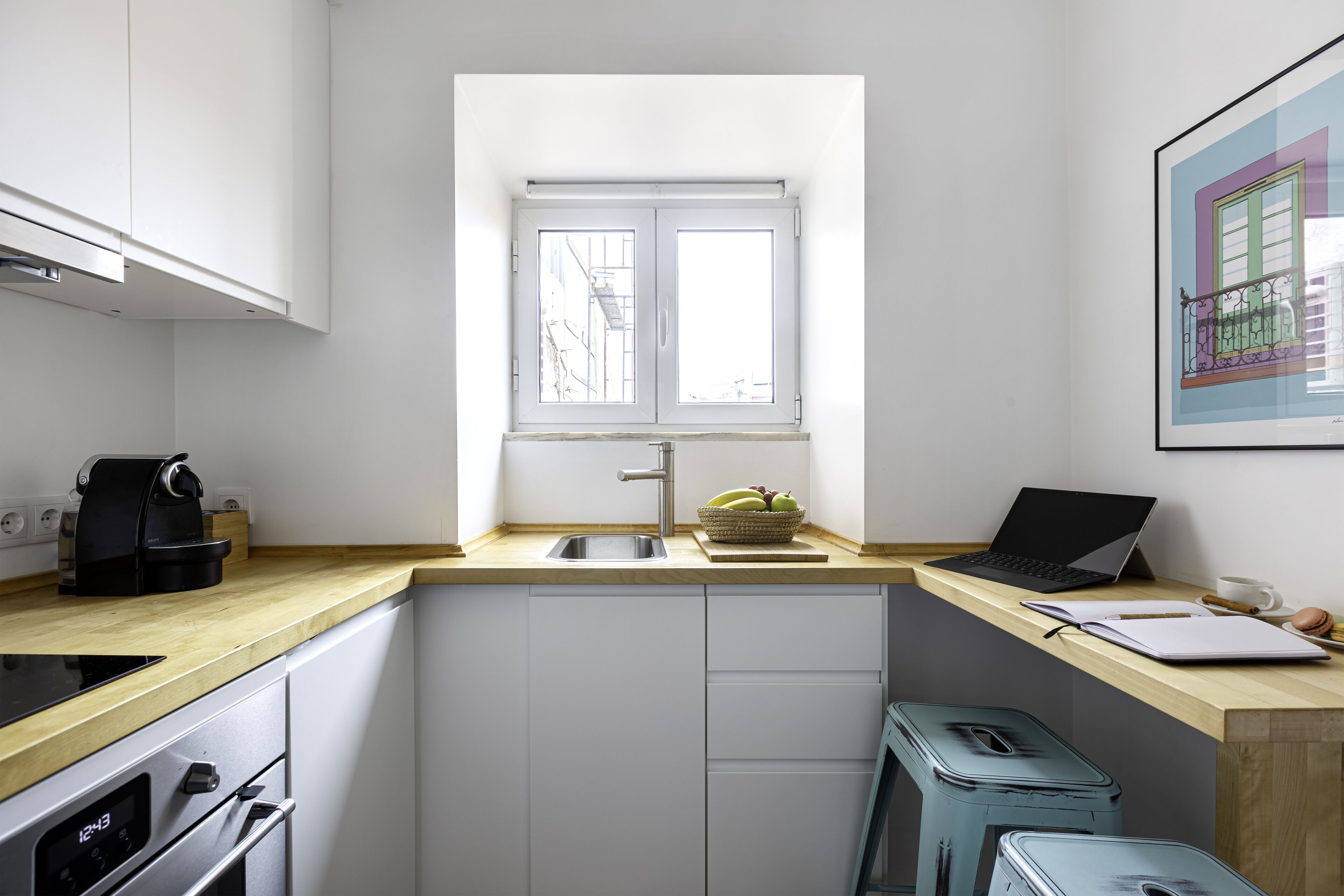 A compact kitchen with a window, a sink, a coffee maker, and a laptop on the counter.