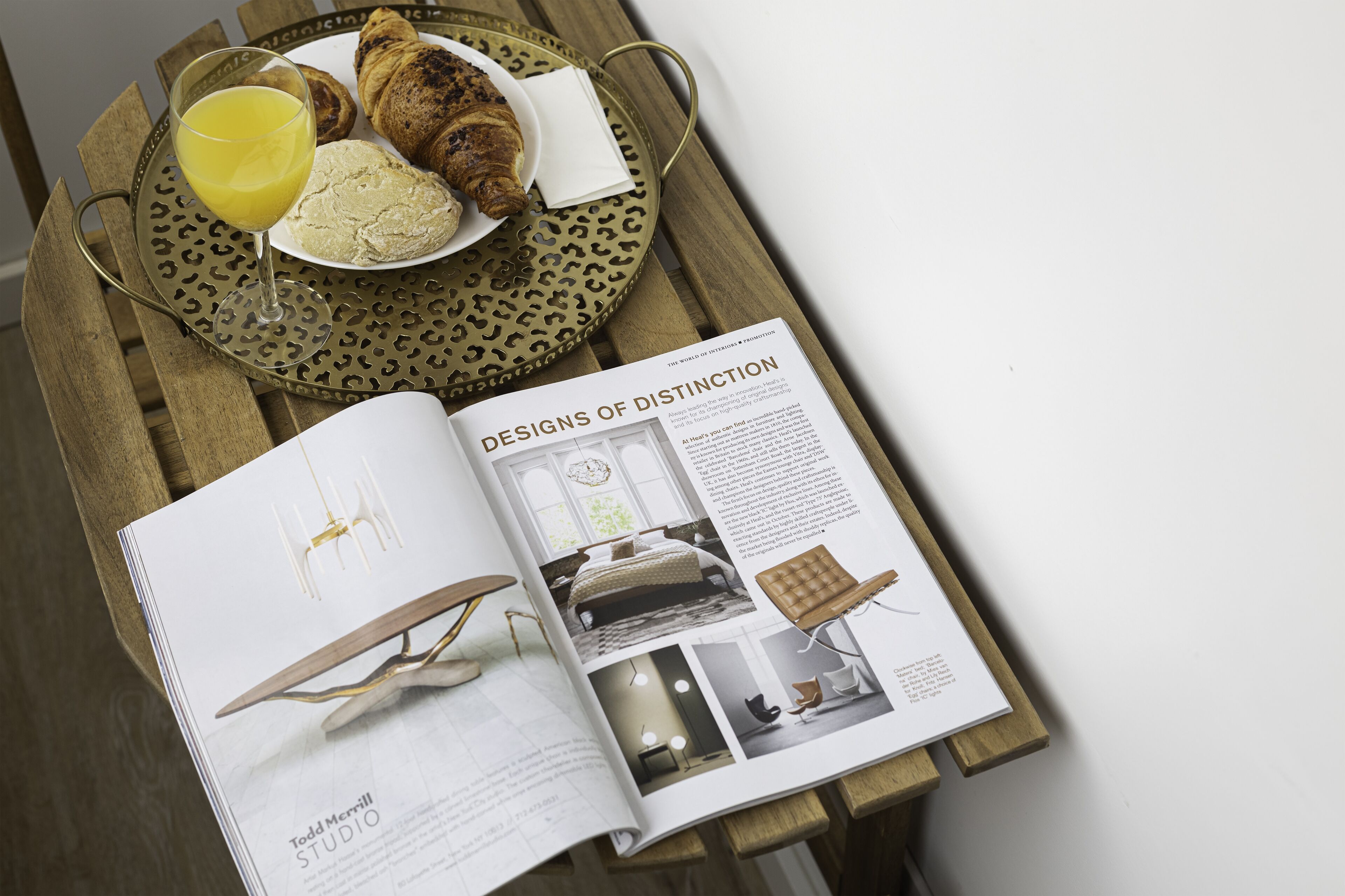 A magazine open to a page titled "DESIGNS OF DISTINCTION" with a table setting that includes croissants, a glass of orange juice, and a napkin.