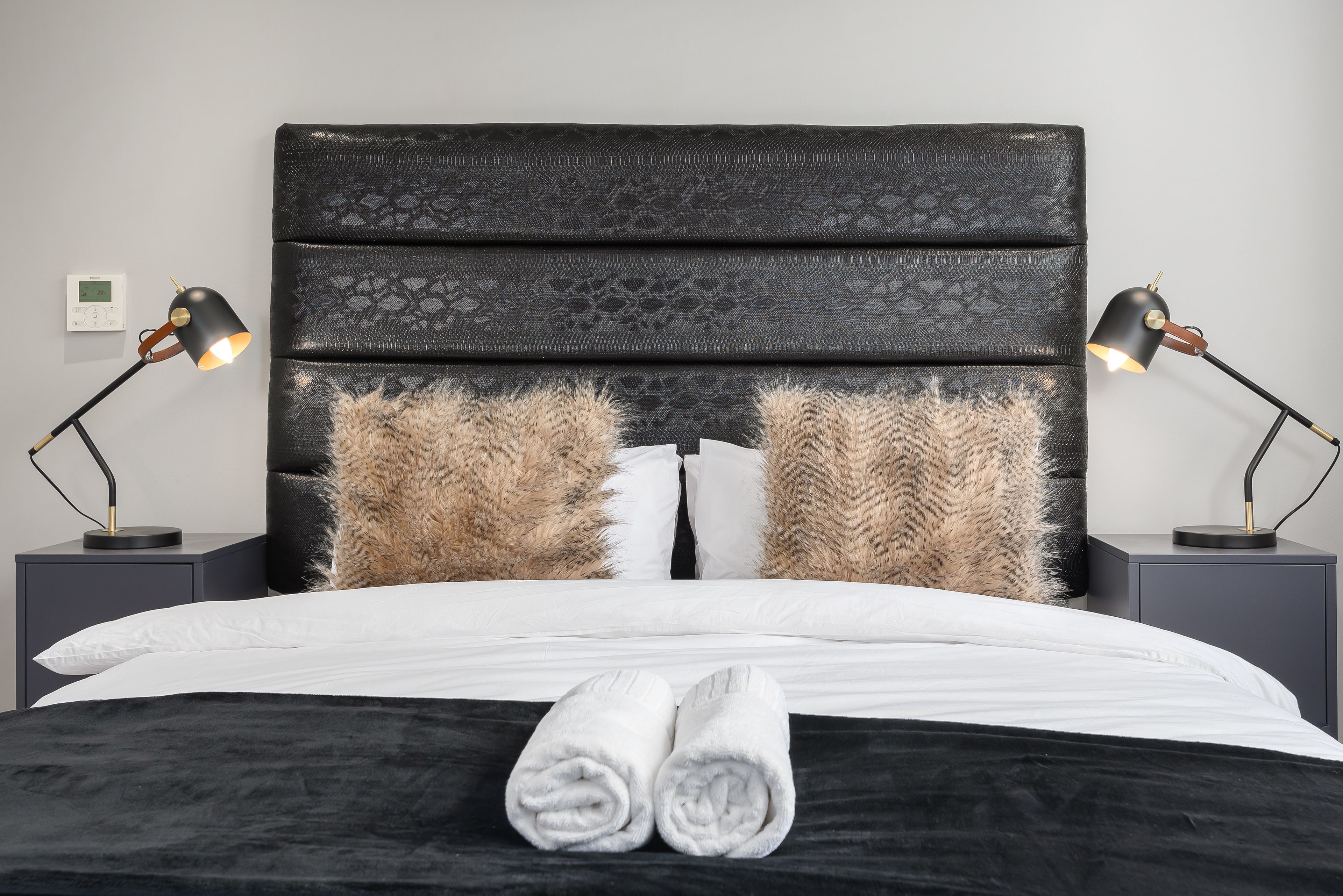 A neatly made bed with a black headboard, two fluffy pillows, and a pair of rolled towels on the bed.