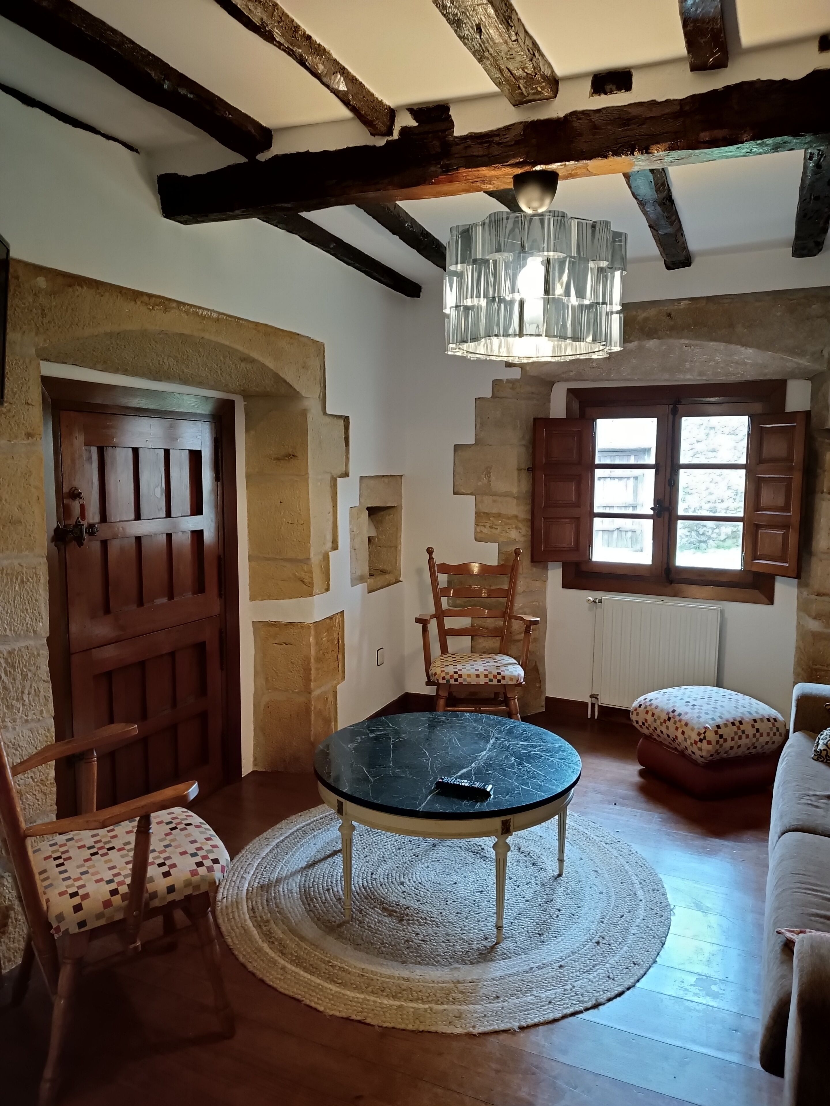 Una habitación rústica con vigas de madera a la vista, una mesa redonda con sillas, una araña y una ventana con contraventanas.