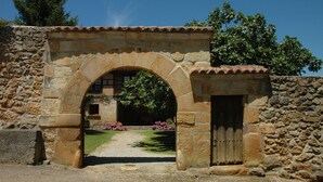 Un portail en pierre en forme d’arc donne accès à un bâtiment doté d’une porte en bois, le tout étant entouré d’un mur de pierre et de verdure.